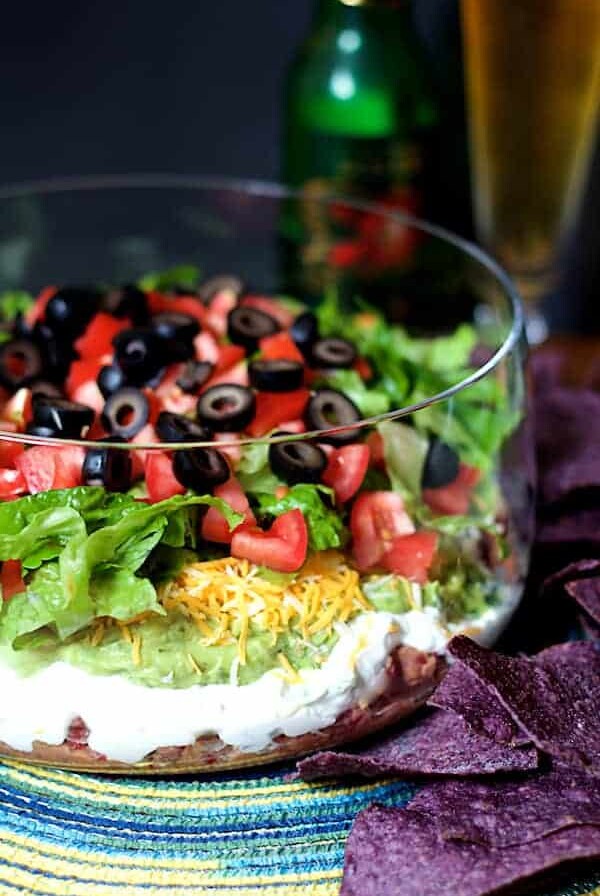Healthier 7-Layer Dip in tall glass bowl with blue corn chips and beer in the background.