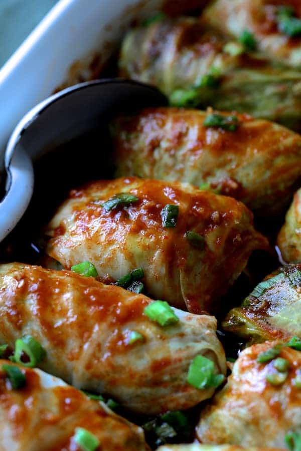 Close-up photo of cooked Spicy Asian Pork Cabbage Rolls in white baking dish with white metal spoon.