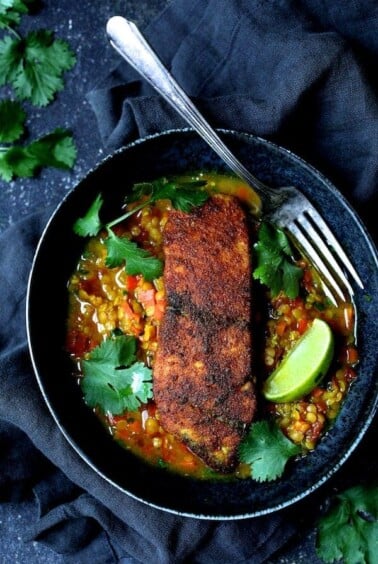 Indian Spiced Sea Bass with Braised Red Lentils in black bowl with fork, cilantro sprigs and lime wedges.