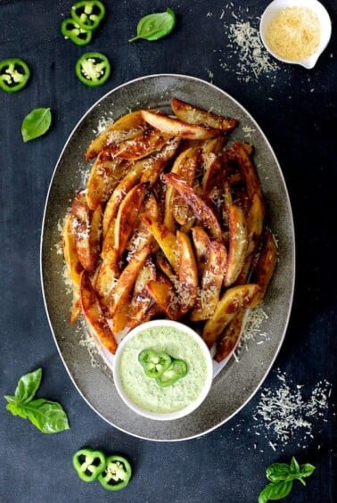 Oven Fries with Spicy Pesto Aioli on gray-rimmed platter garnished with jalapeno slices, fresh basil leaves and parmesan cheese.