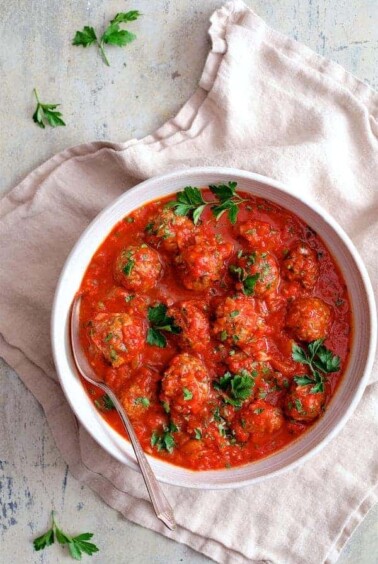 Spiced Meatballs with Tomato Sauce in white bowl with serving spoon on beige napkin.