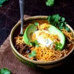 Beef and Chorizo Chili in brown bowl on brown burlap garnished with cheese, sour cream and avocado wedges.