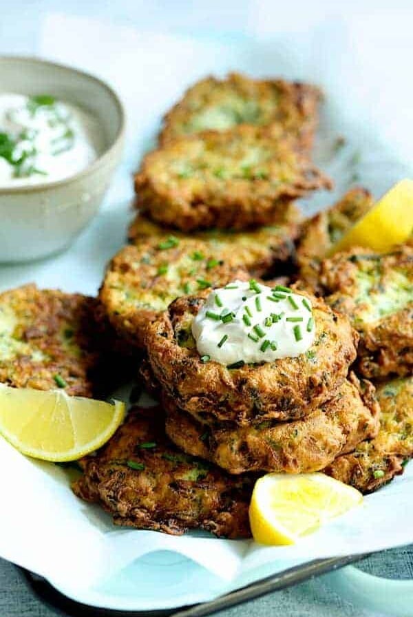 Stacks of Zucchini Ricotta Fritters on platter with yogurt chive topping garnished with lemon slices.