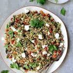 Mediterranean Farro Salad on round white platter with serving utensils garnished with parsley.