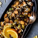 Pan-Roasted Brussels Sprouts with Thai Sweet Chili Orange Glaze on black oval platter with serving fork garnished with almonds and orange wedges.