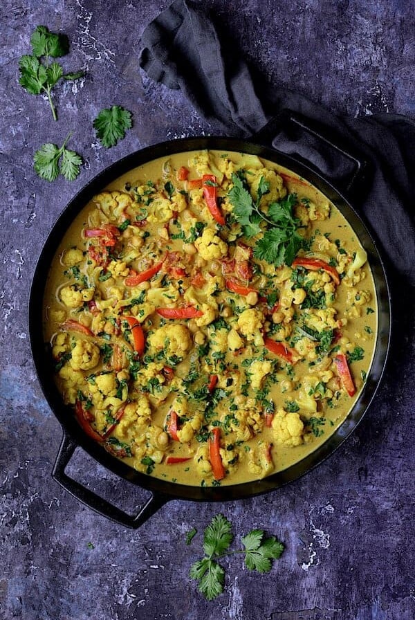 Vegan Indian Cauliflower and Chickpea Curry - Overhead shot of dish in cast iron pan on blue background with gray towel garnished with cilantro