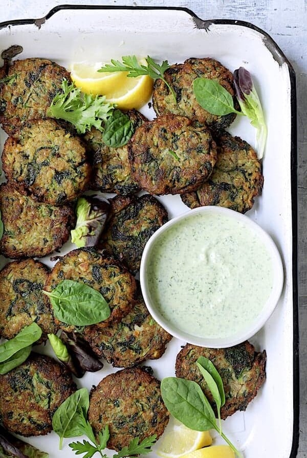 Zucchini Red Lentil Fritters with Cilantro Yogurt Sauce - Overhead shot in white pan with sauce garnished with greens and lemons