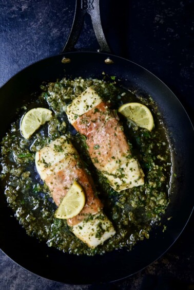 Photo of Prosciutto-Wrapped Halibut with Herb - Caper Butter in black skillet with lemon wedges.