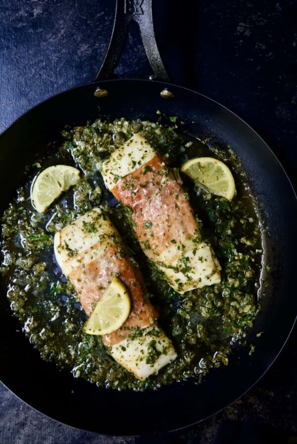 Photo of Prosciutto-Wrapped Halibut with Herb - Caper Butter in black skillet with lemon wedges.