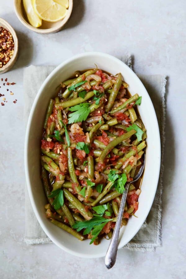 Mediterranean Braised Green Beans with Tomatoes in oval serving dish with serving spoon garnished with fresh parsley.