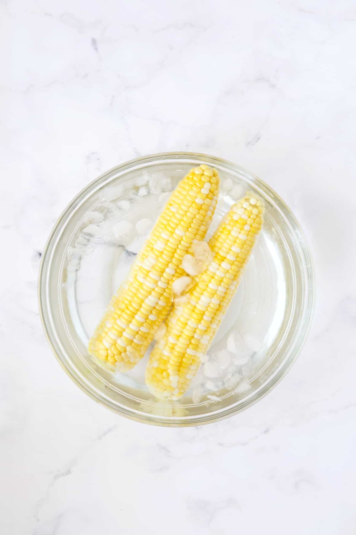 Two cobs of corn in ice water in glass bowl.