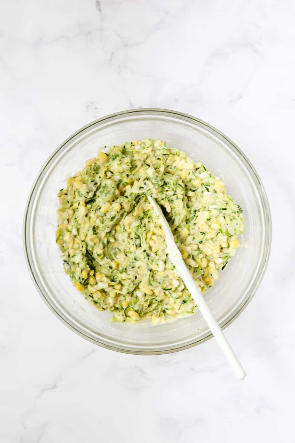 Batter for Zucchini Corn Fritters in glass bowl being stirred with white spoon.