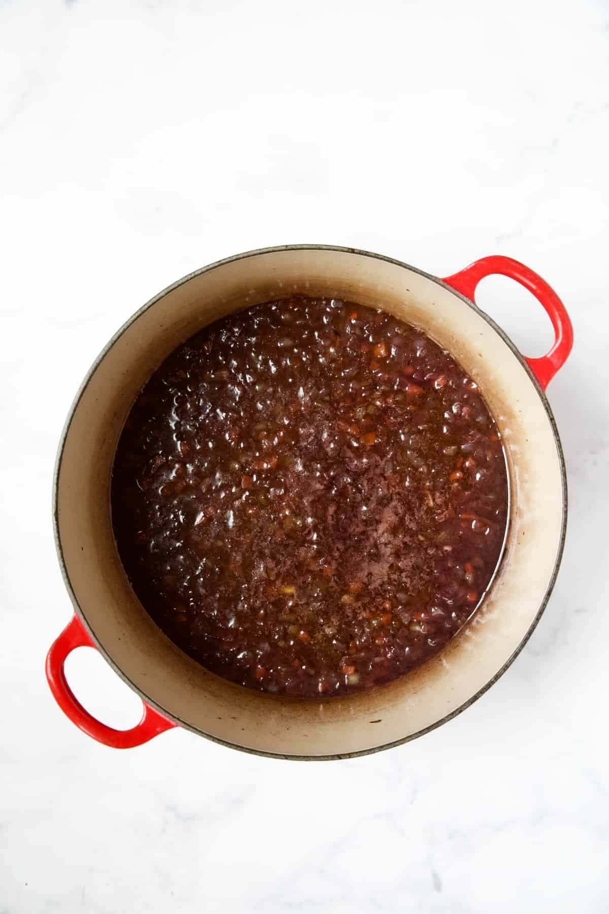 Red wine added to vegetables in red enamel Dutch oven.