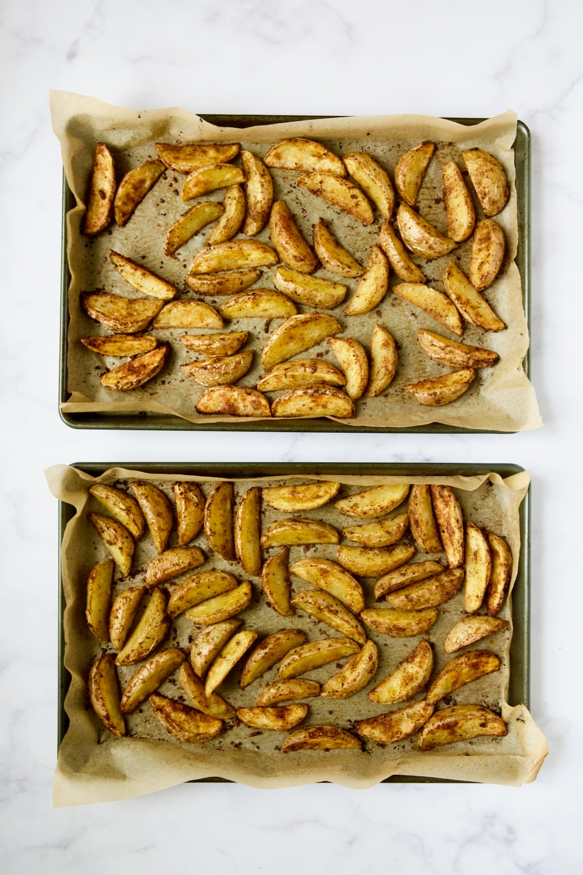 Two baking sheets lined with parchment paper of roasted potato wedges.