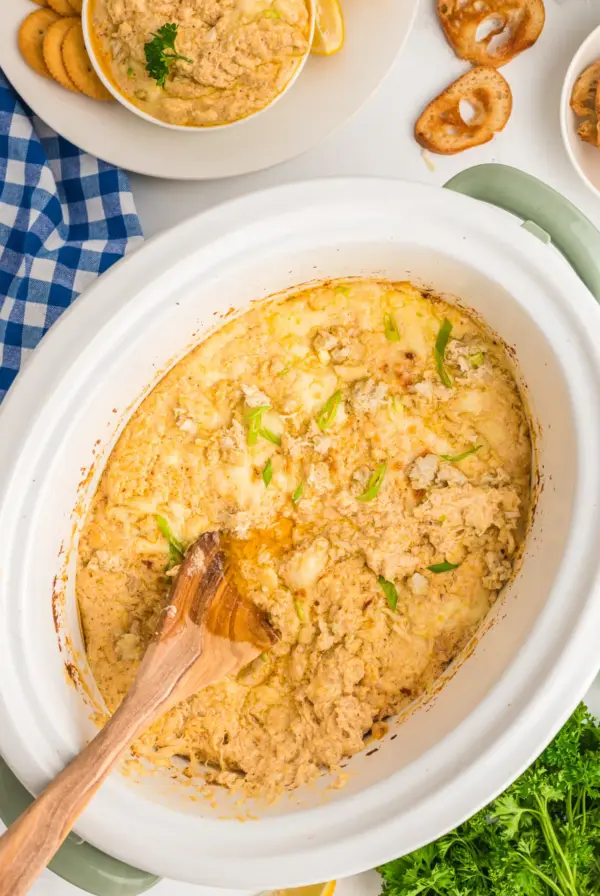 Crab Dip in white slow cooker with wooden spoon stirring it.