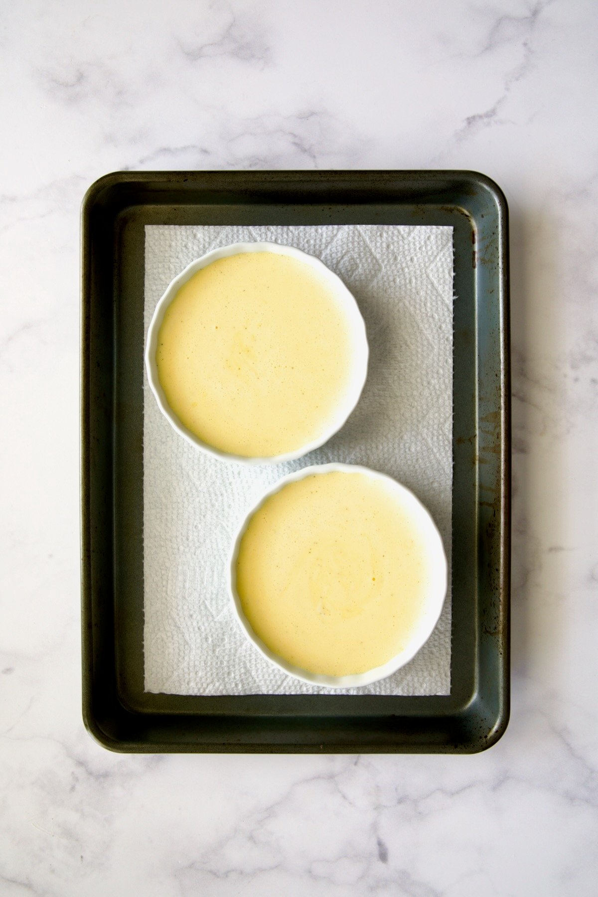 Creme brulee batter poured into white creme brulee dishes set on paper towel in rimmed sheet pan.