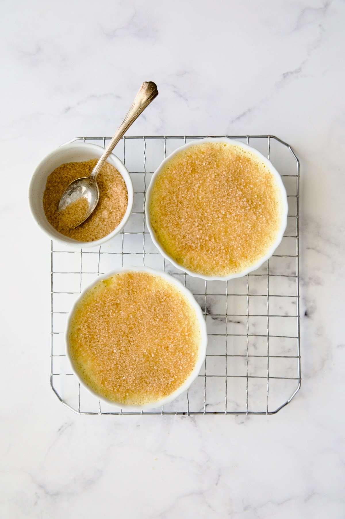 Two creme brulees on wire rack with turbinado sugar sprinkled over the top with bowl of turbinado. sugar