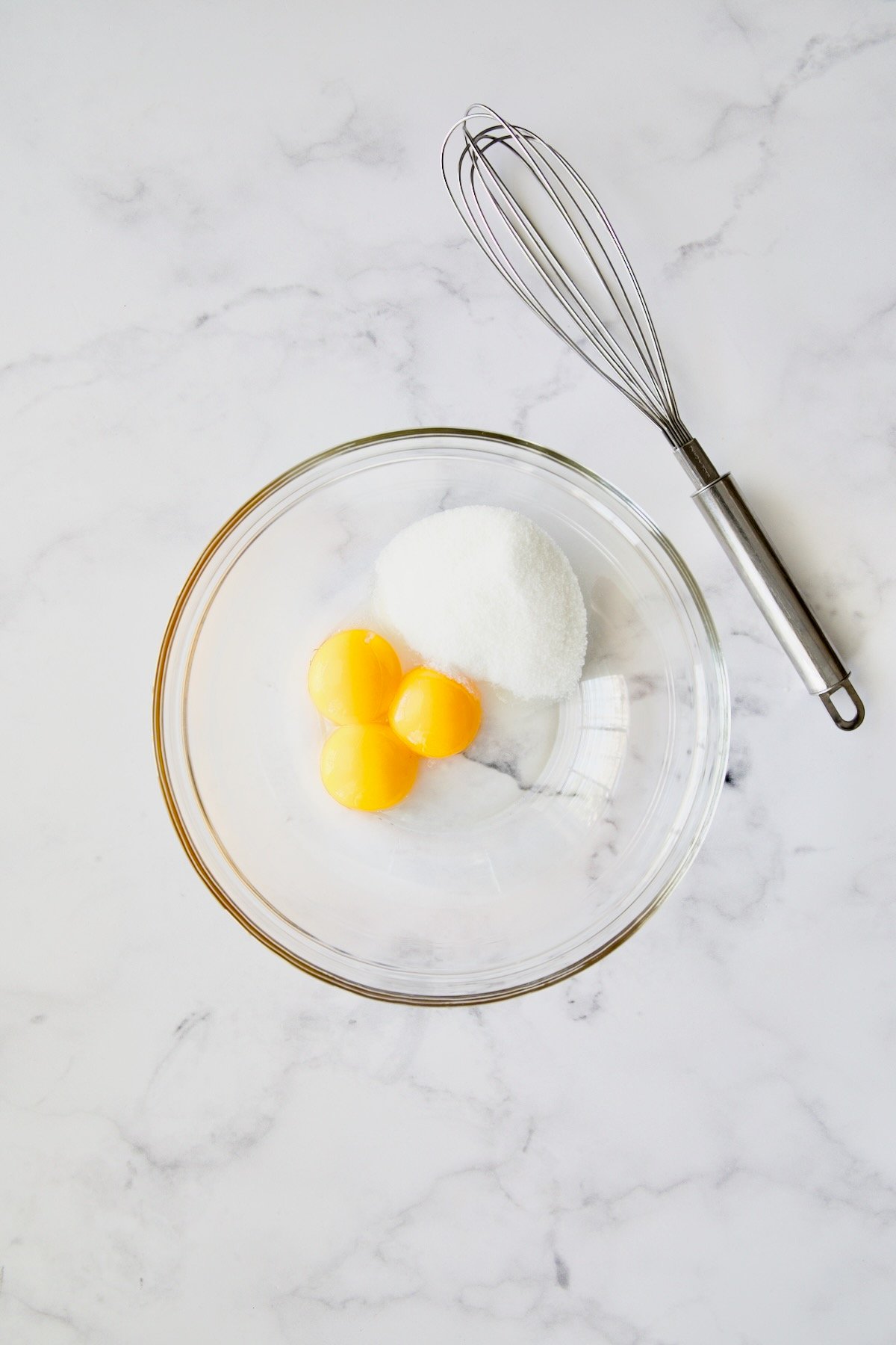 Three egg yolks and granulated sugar in glass bowl with wire whisk.