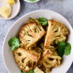 Four wedges of Curry Roasted Cauliflower Wedges with Cilantro Chutney in white bowl with lemon wedges.