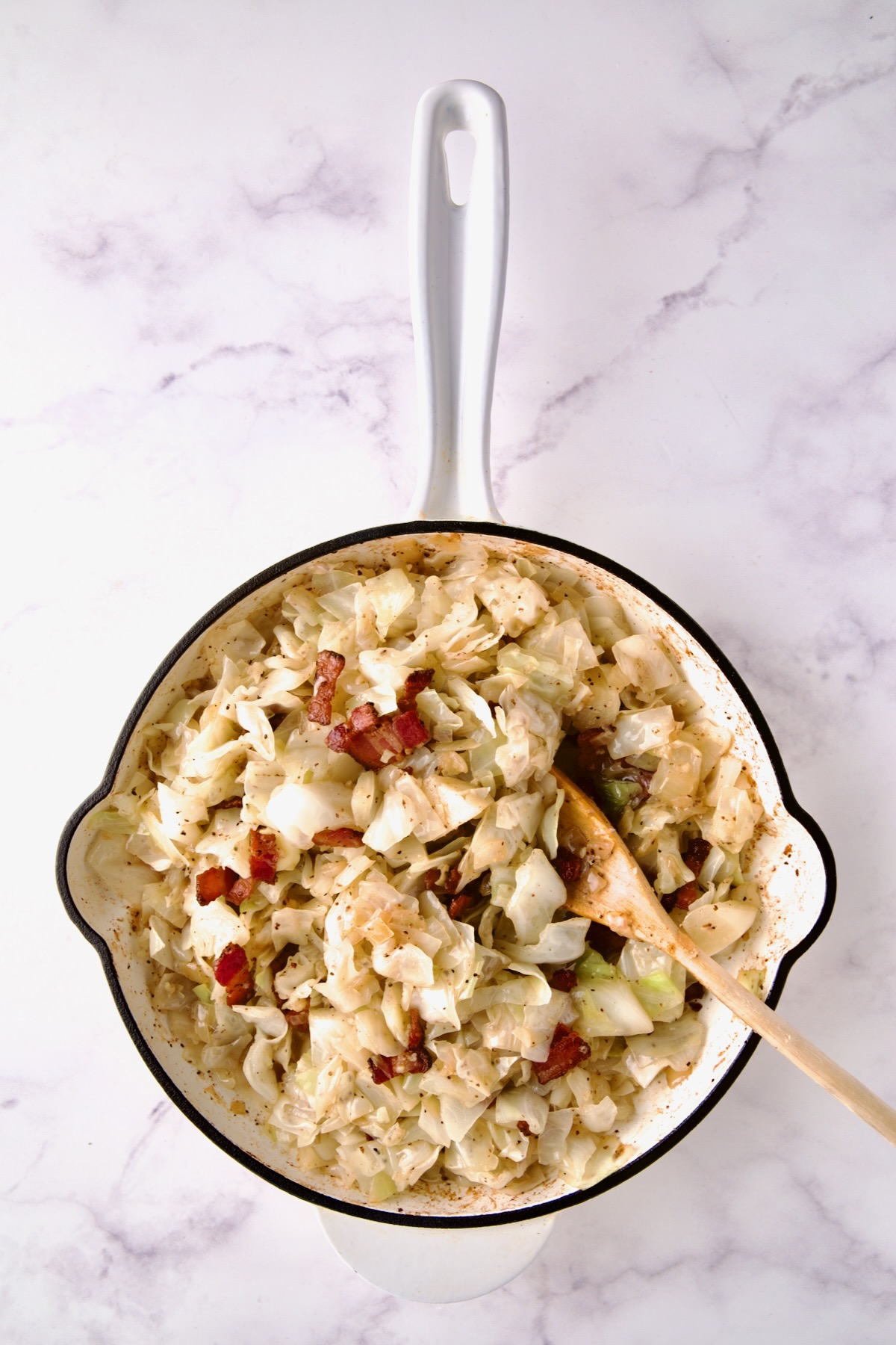 Bacon being stirred back into cabbage in white enamel cast iron skillet.