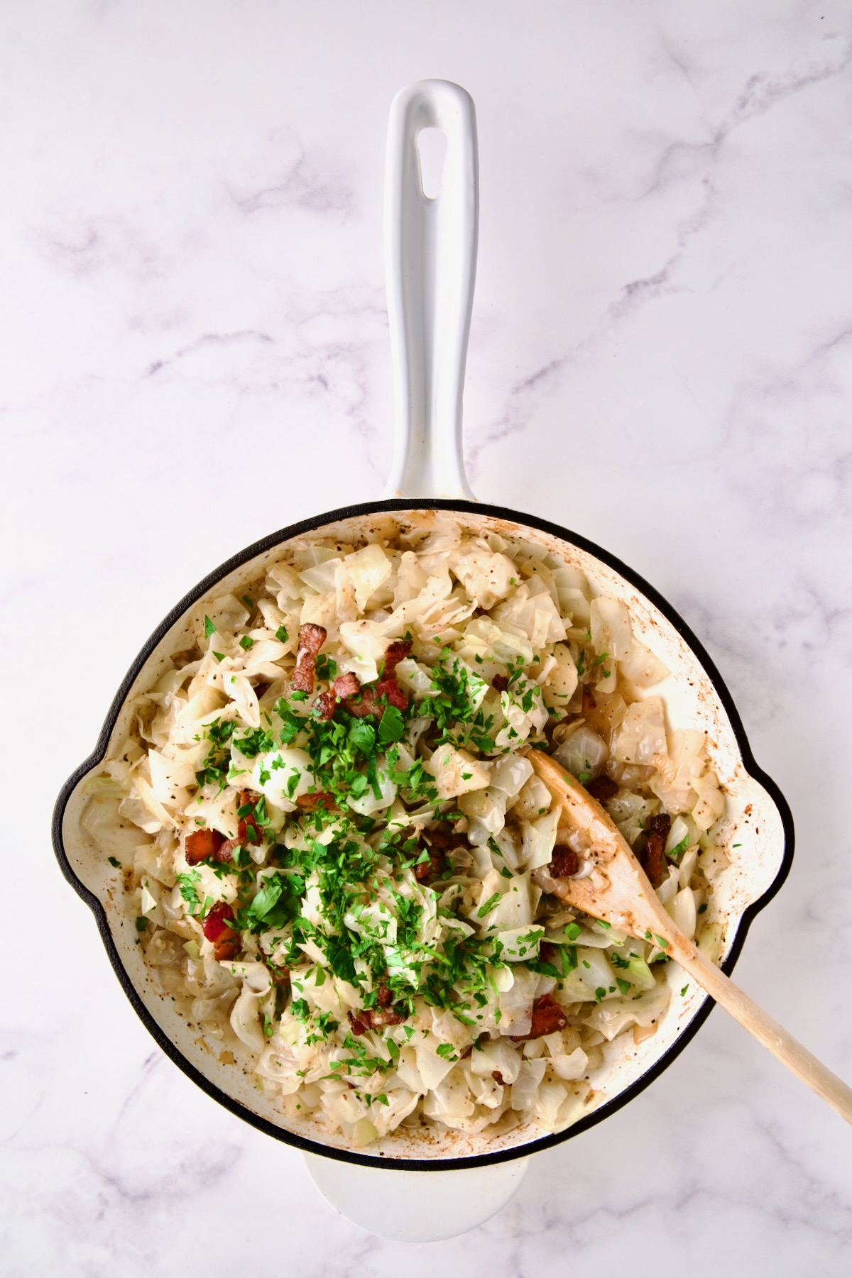 Cabbage in white enamel cast iron skillet with bacon and parsley added being stirred with wooden spoon.
