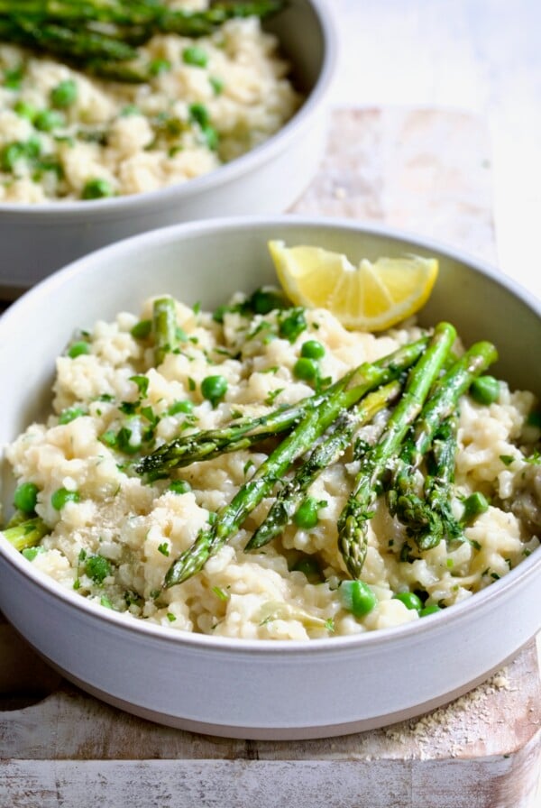 One bowl of Easy Risotto Recipe with Spring Vegetables with asparagus spears on the top and lemon wedge on whitewashed cutting board.