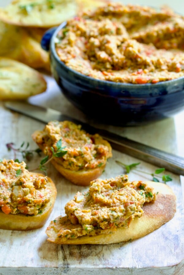 Three pieces of crostini topped with Artichoke Tapenade with bowl of tapenade in the background.