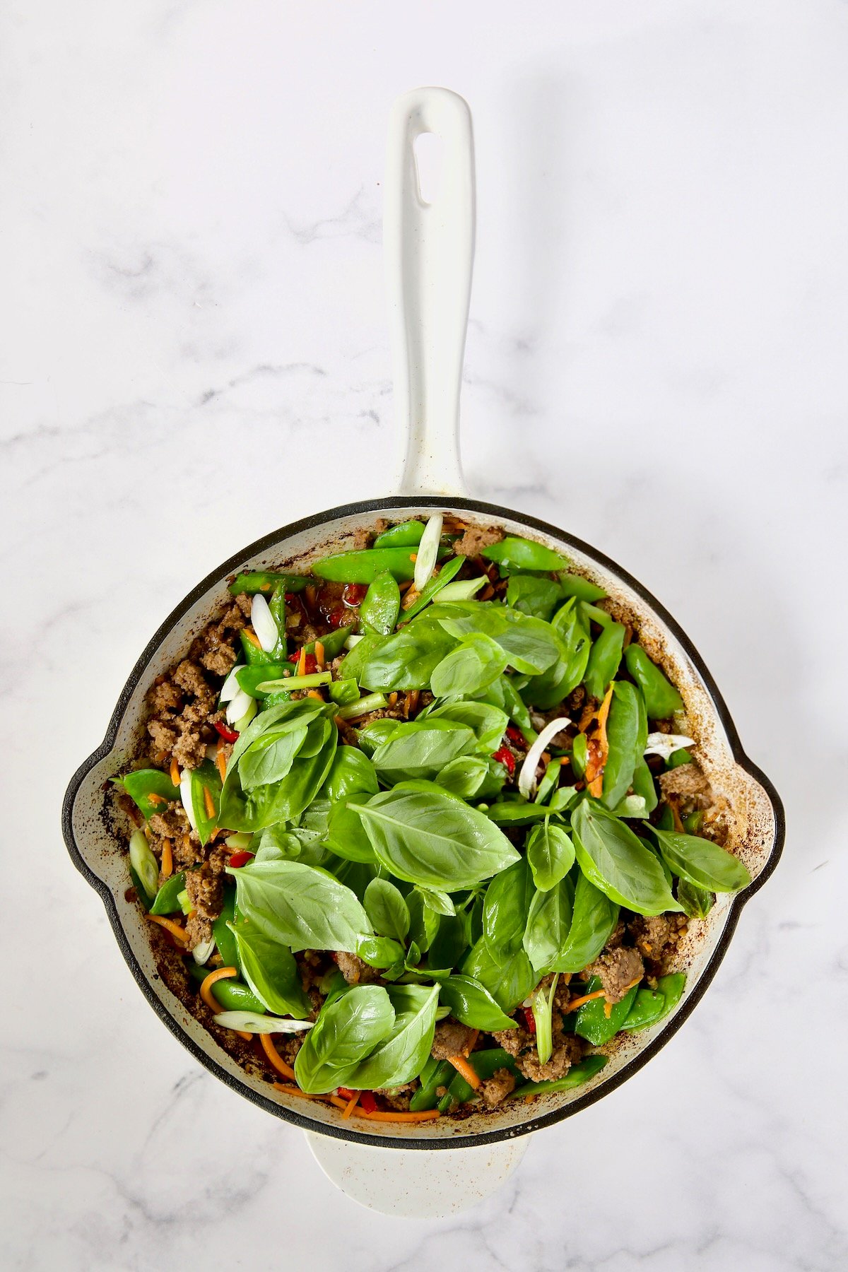 Cooked ground beef, Fresno chiles, shredded carrots, snow peas, scallions and fresh basil in white enamel cast iron skillet.