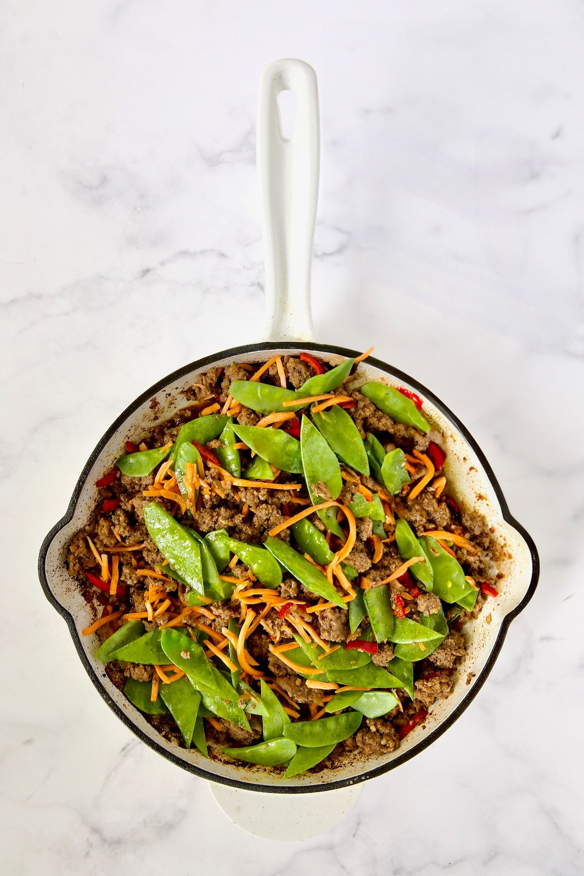 Cooked ground beef, Fresno chiles, shredded carrots and snow peas in white enamel cast iron skillet.