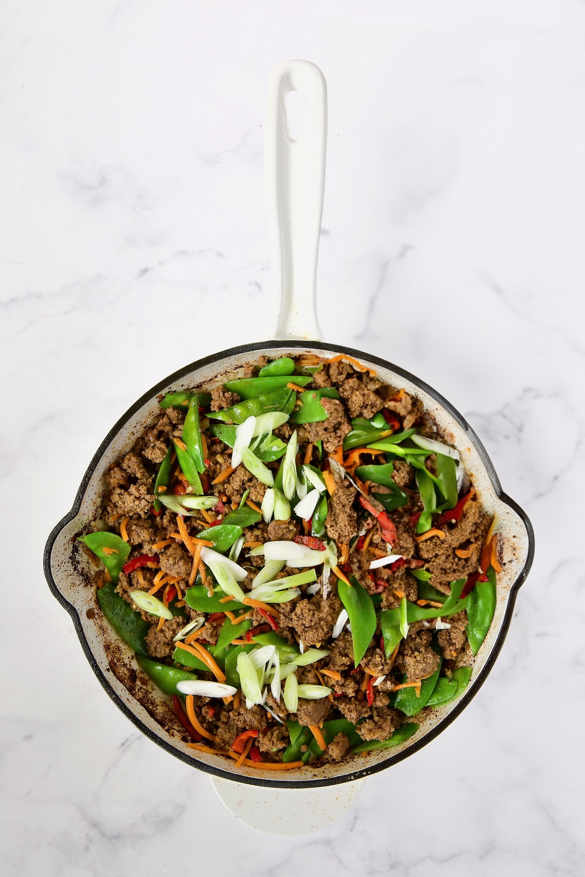 Cooked ground beef, Fresno chiles, shredded carrots, snow peas and scallions in white enamel cast iron skillet.