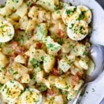 German Potato Salad in round earthenware bowl garnished with egg slices and fresh parsley with serving spoon.
