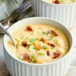 Cheddar Bacon Potato Soup in large white soup mug with spoon garnished with bacon, scallions and cheese shreds.