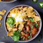 One bowl of Dutch Oven Chili with corn chips, cheese, sour cream jalapeno slices and cilantro in gray bowl.