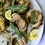Roasted Artichokes with Garlic Butter on white oval platter garnished with lemon wedges and herb sprigs.