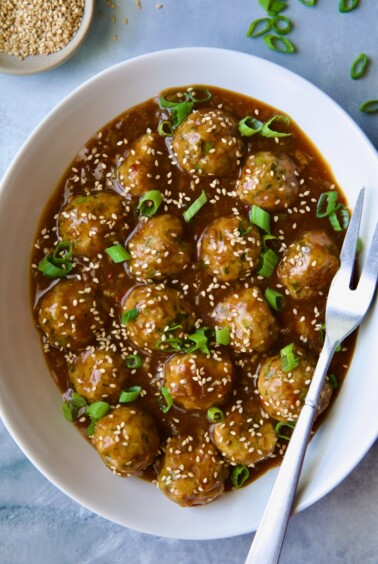 Asian Turkey Meatballs in white oval serving bowl with serving fork garnished with sesame seeds and scallions.