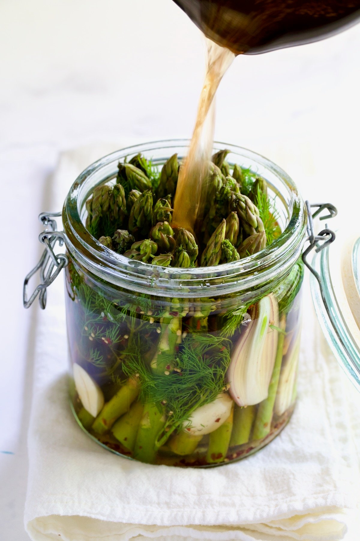 Brine being poured over asparagus, garlic, shallot and dill in glass jar.