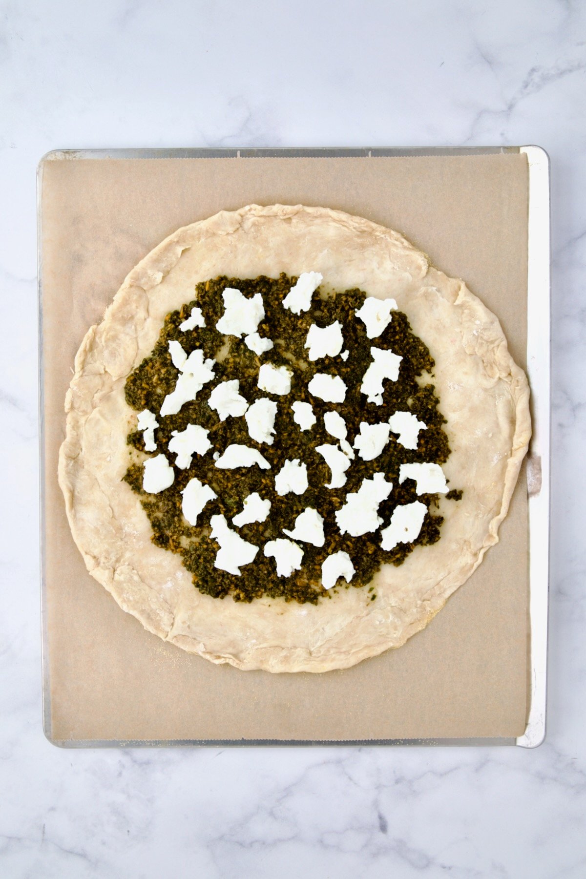 Rolled out galette dough on parchment paper on cookie sheet with pesto and goat cheese added.