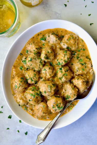 Baked Turkey Meatballs in Beer - Onion Sauce in white oval bowl with serving spoon garnished with fresh parsley.