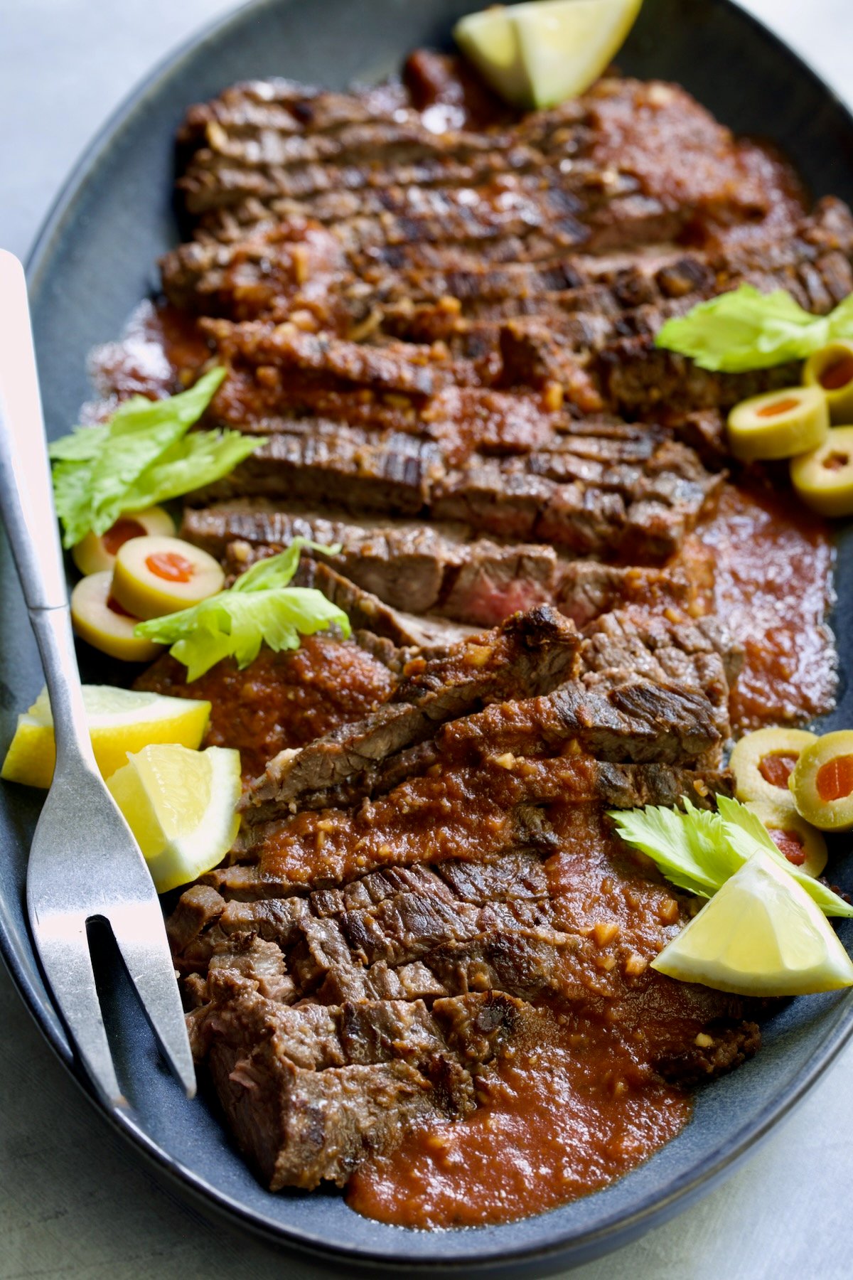 Sliced Bloody Mary Skirt Steak on blue oval platter garnished with lemon wedges, olive slices and celery leaves.