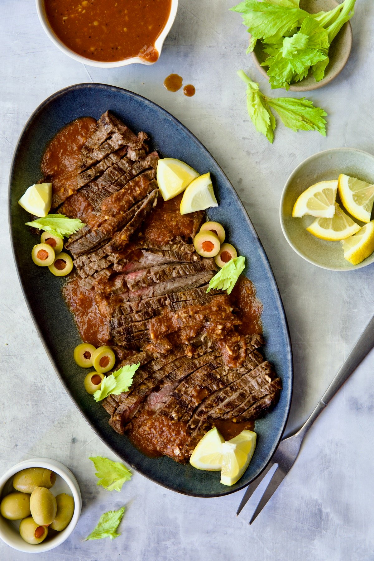 Sliced Bloody Mary Skirt Steak on blue oval platter garnished with lemon wedges, olive slices and celery leaves.