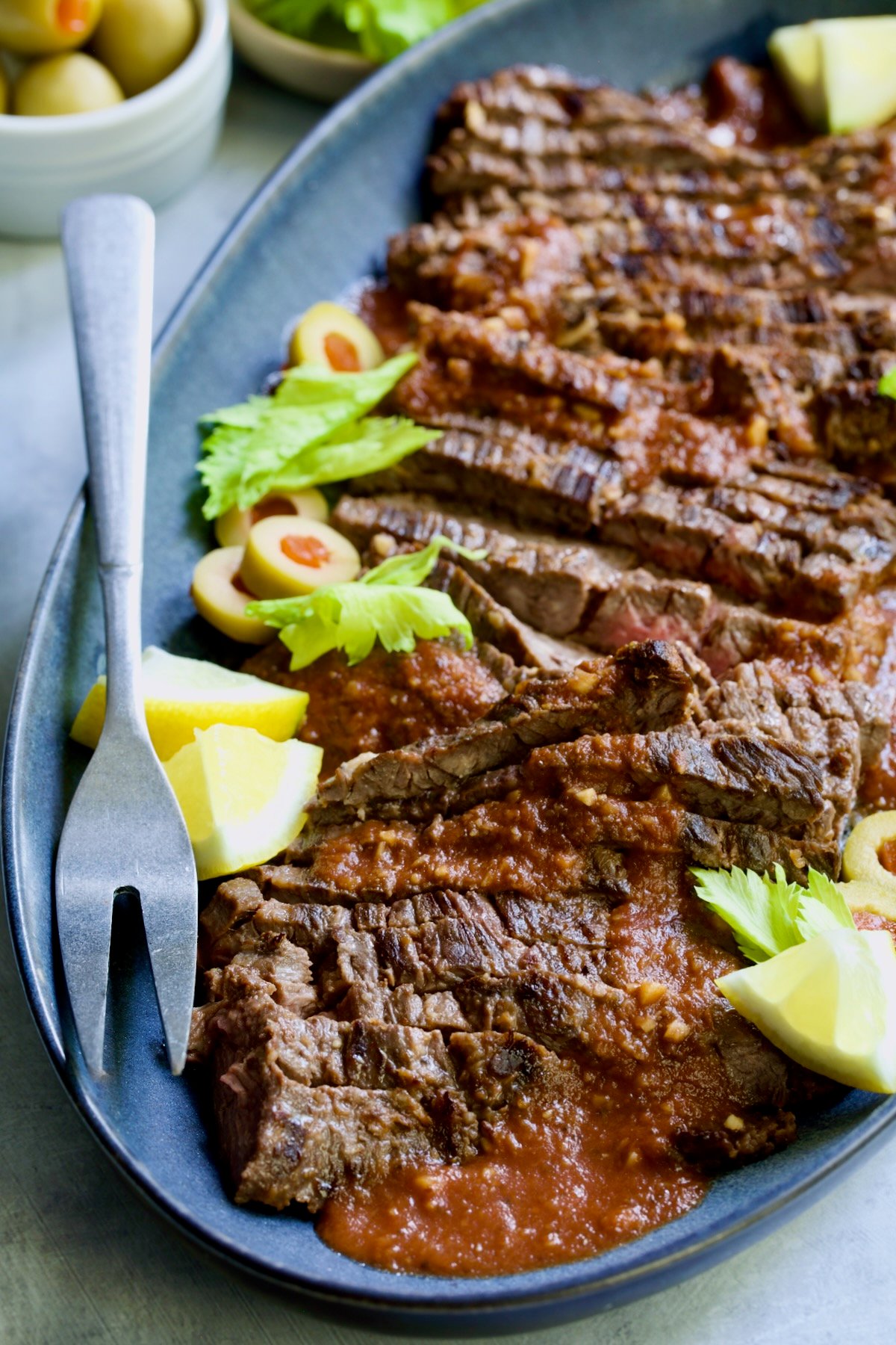 Sliced Bloody Mary Skirt Steak on blue oval platter garnished with lemon wedges, olive slices and celery leaves.