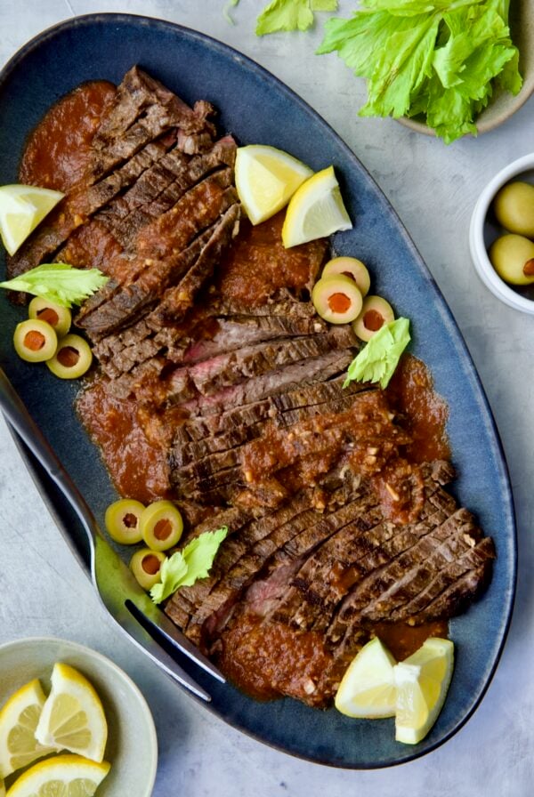 Sliced Bloody Mary Skirt Steak on blue oval platter garnished with lemon wedges, olive slices and celery leaves.