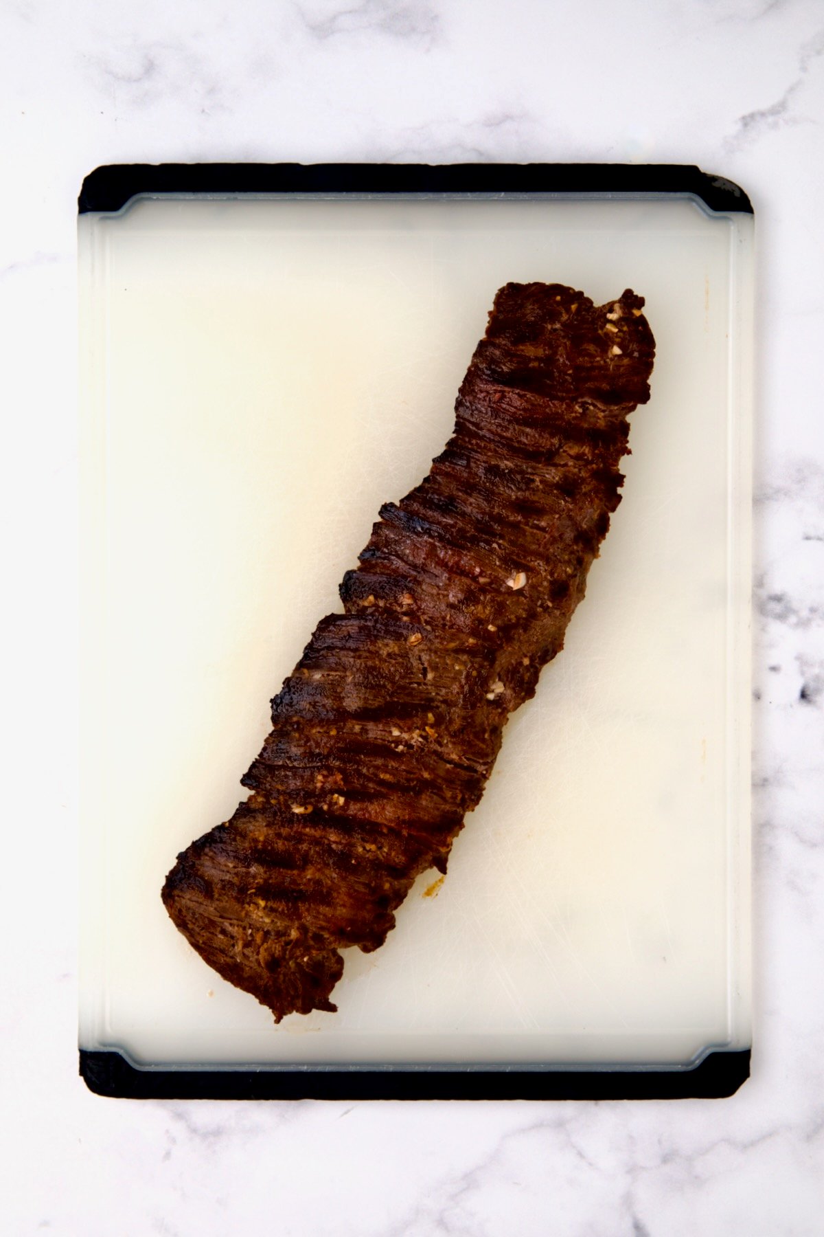 Grilled skirt steak on white plastic cutting board before being sliced.