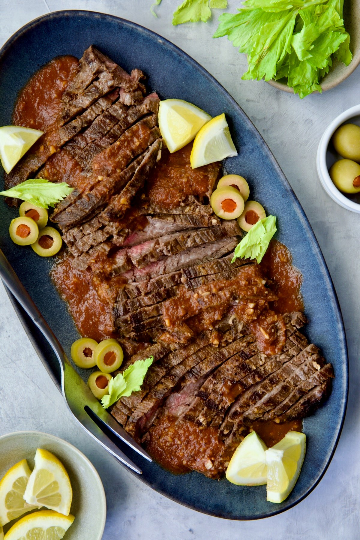 Sliced Bloody Mary Skirt Steak on blue oval platter garnished with lemon wedges, olive slices and celery leaves.