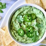 Spicy Guacamole in white bowl surrounded by tortilla chips garnished with jalapeno slices and cilantro leaves.