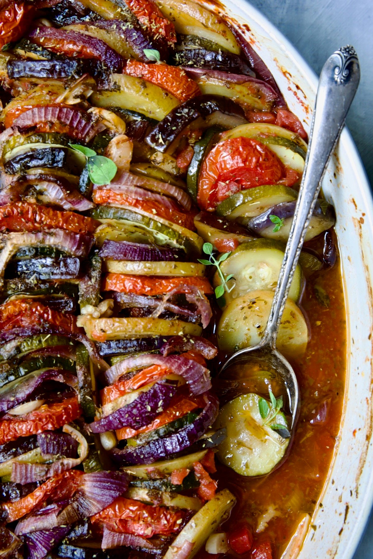 Briam (Greek Roast Vegetables) in oval white gratin dish with serving spoon exposing the tomato and olive oil juices.