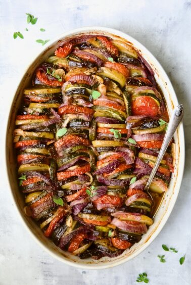Briam (Greek Roast Vegetables) in oval white gratin dish with serving spoon garnished with fresh oregano leaves.