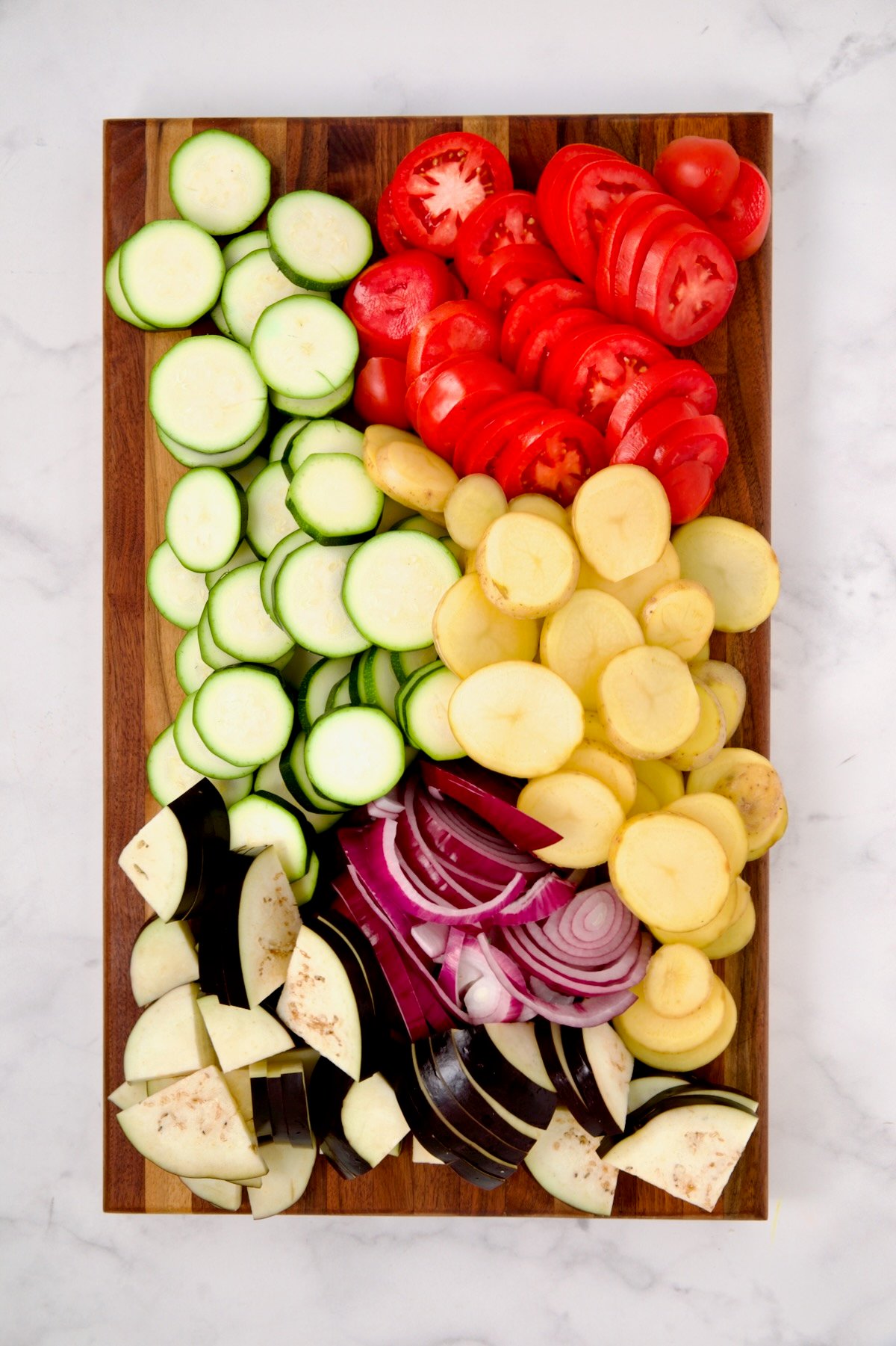Sliced zucchini, potatoes, tomatoes, eggplant and red onion on wood cutting board.