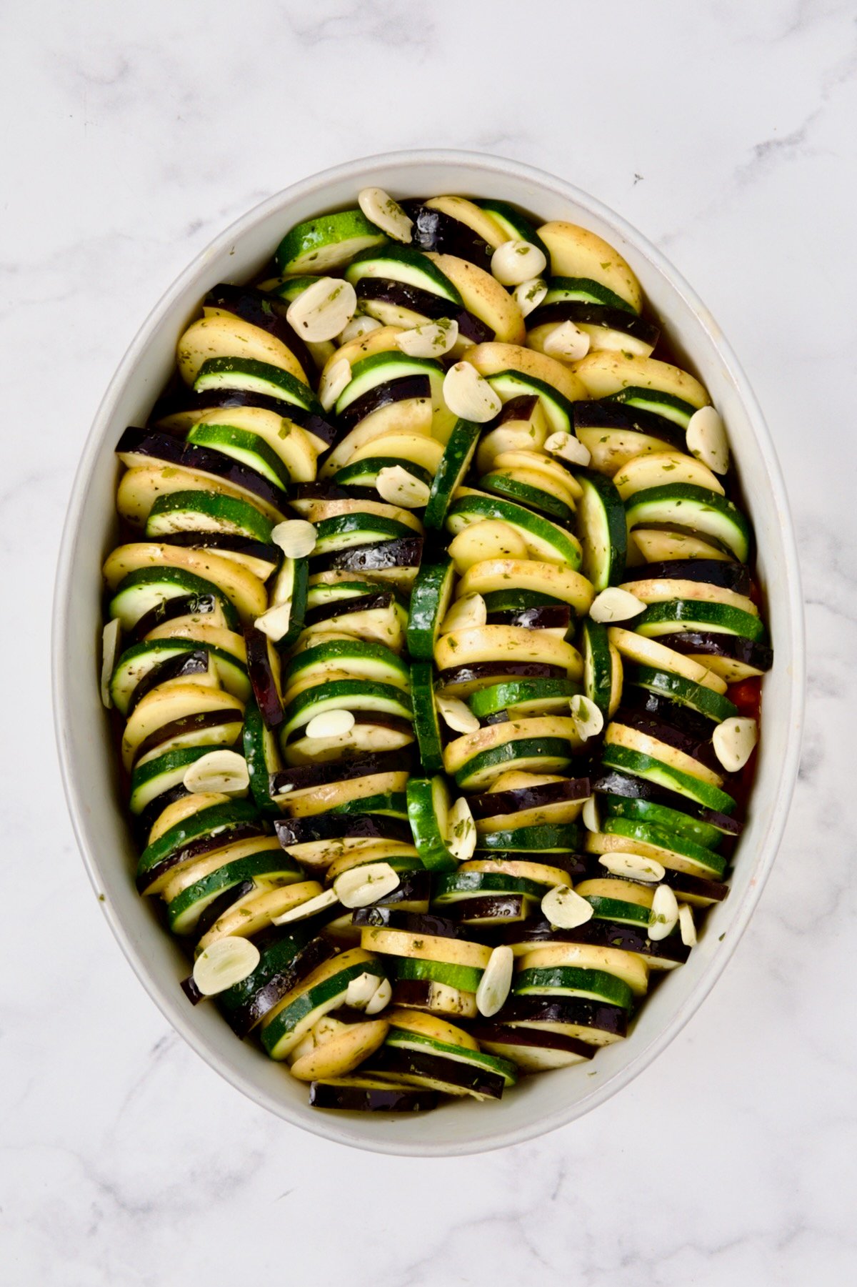Potatoes, zucchini and eggplant arranged in oval white baking dish with sliced garlic over the top.