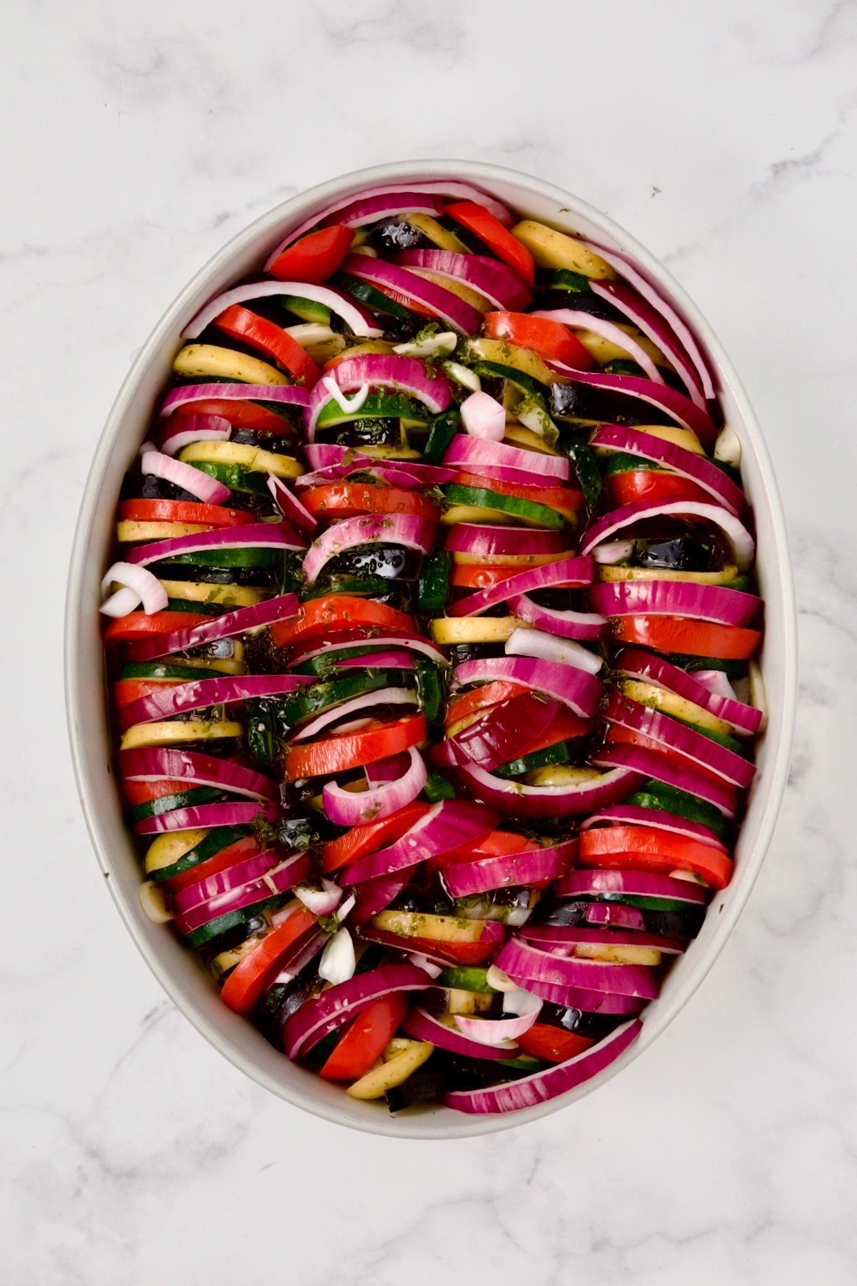 Red onion and tomatoes added to potatoes, zucchini and eggplant in oval white baking dish ready to bake.